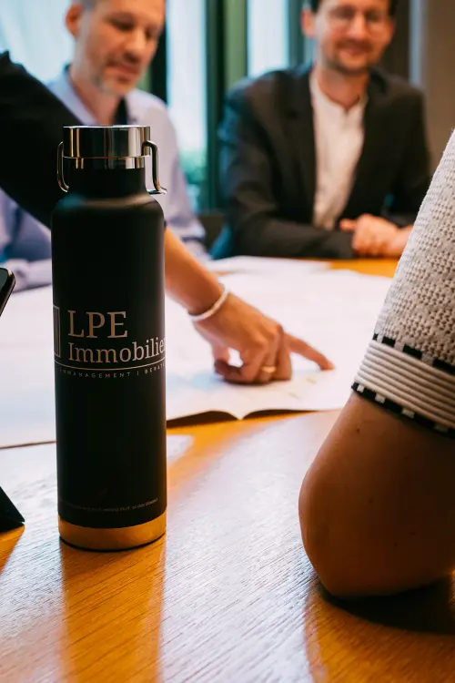 Besprechung am Tisch – Hand zeigt auf Bauplan, LPE-Immobilien Flasche im Vordergrund