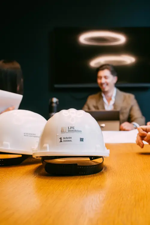 Weißer Schutzhelm mit LPE-Logo im Vordergrund, Besprechung im Hintergrund unscharf