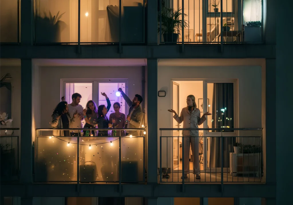 Nacht: links Balkonparty mit sechs Personen und Lichterkette, rechts genervte Nachbarin im Pyjama; modernes Mehrfamilienhaus in Deutschland.