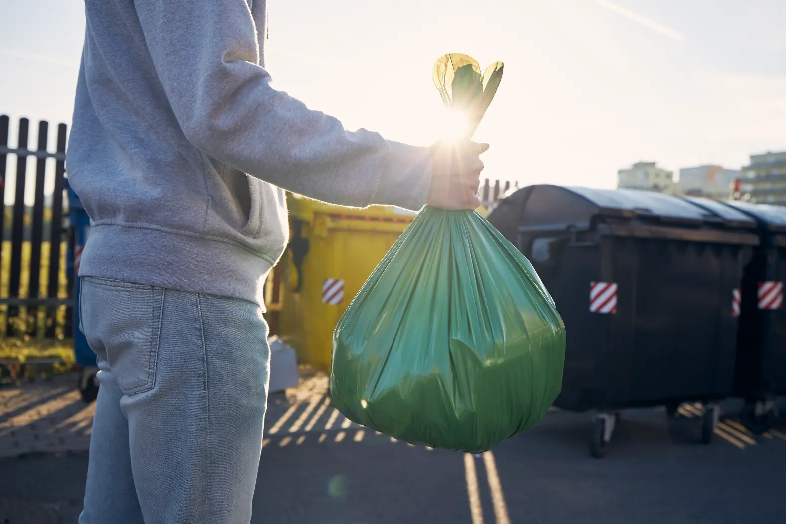 Mann trägt einen verschlossenen grünen Müllsack zu Sammelcontainern auf dem Hof einer Wohnanlage – Symbolfoto für korrekte Mülltrennung in der WEG.