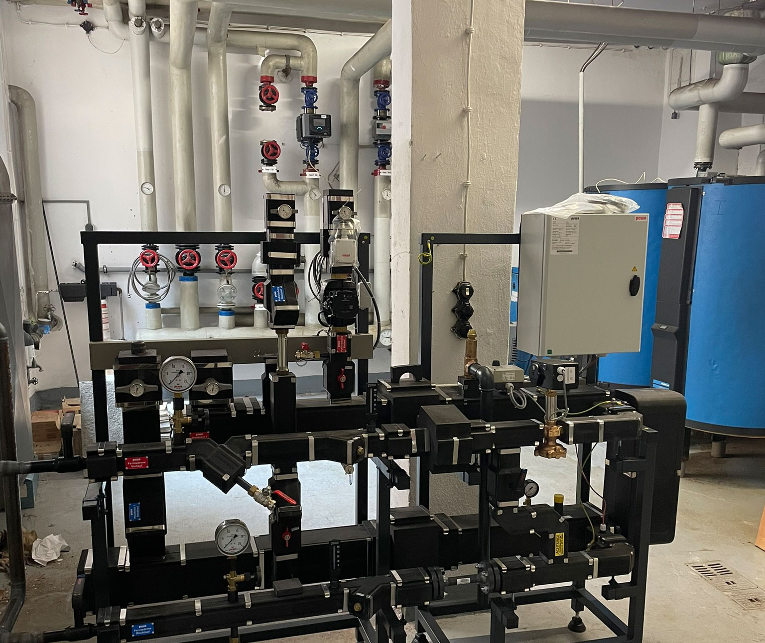 Heizungsraum mit Fernwärme-Übergabestation: schwarze Rahmenkonstruktion mit Pumpen, Ventilen und Manometern vor weißen Rohrleitungen und blauen Speichern.
